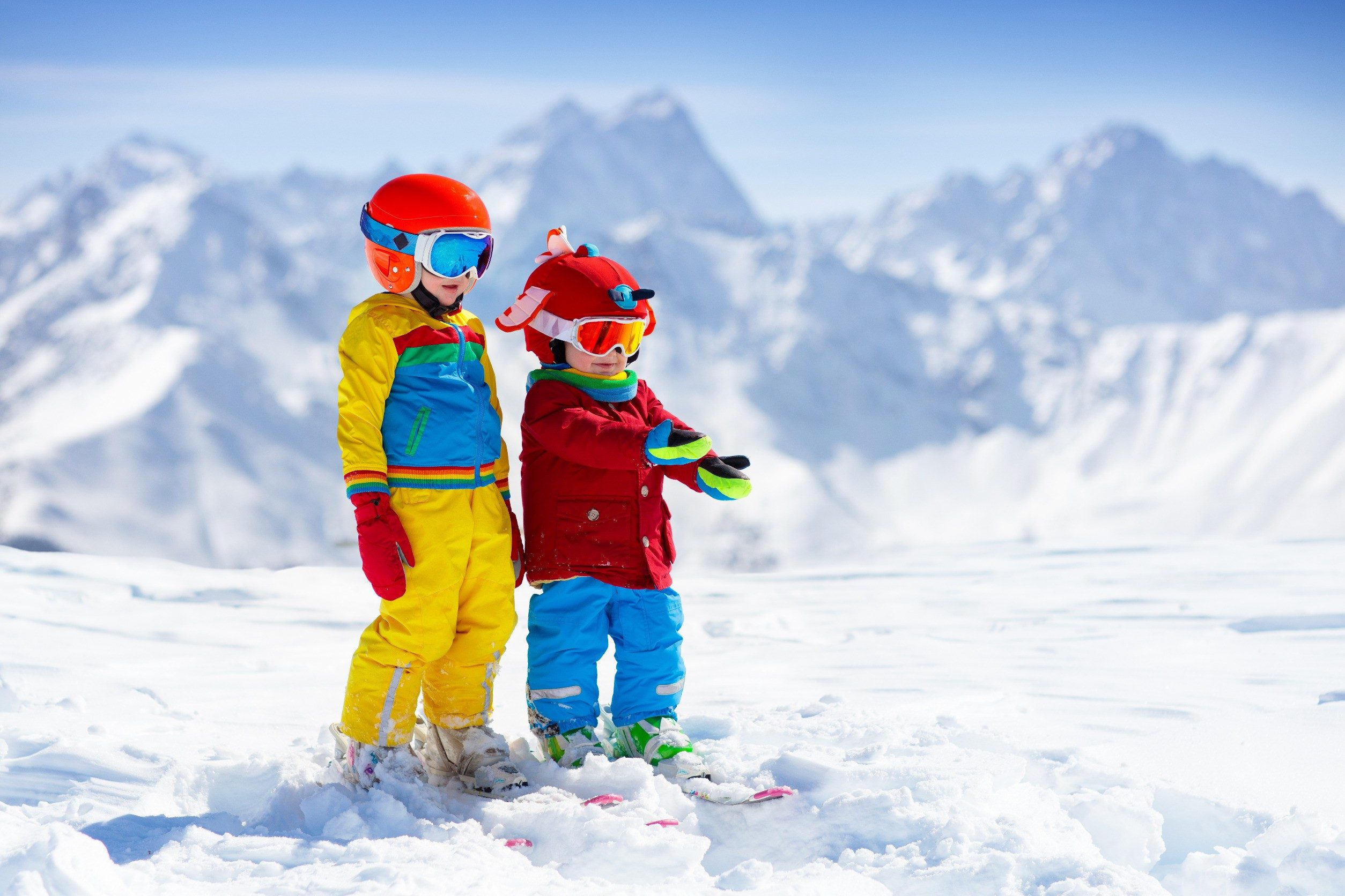 Two young children in bright snowsuits and ski helmets standing in the snow